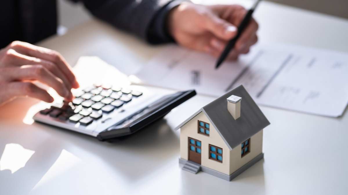 Las manos de una persona haciendo cálculos junto a una figura de una vivienda